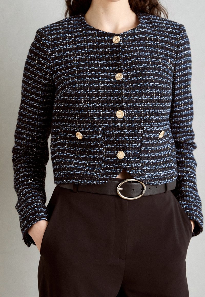Femme portant une veste boutonnée texturée noire et bleue avec des boutons dorés et un pantalon noir avec une ceinture noire avec une boucle ovale argentée.