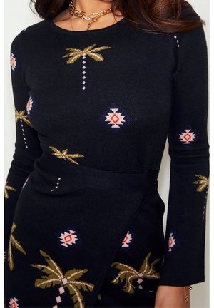 Kvinde iført en sort langærmet kjole med en palme og geometrisk print, accessorized med lagdelte gyldne kæder.