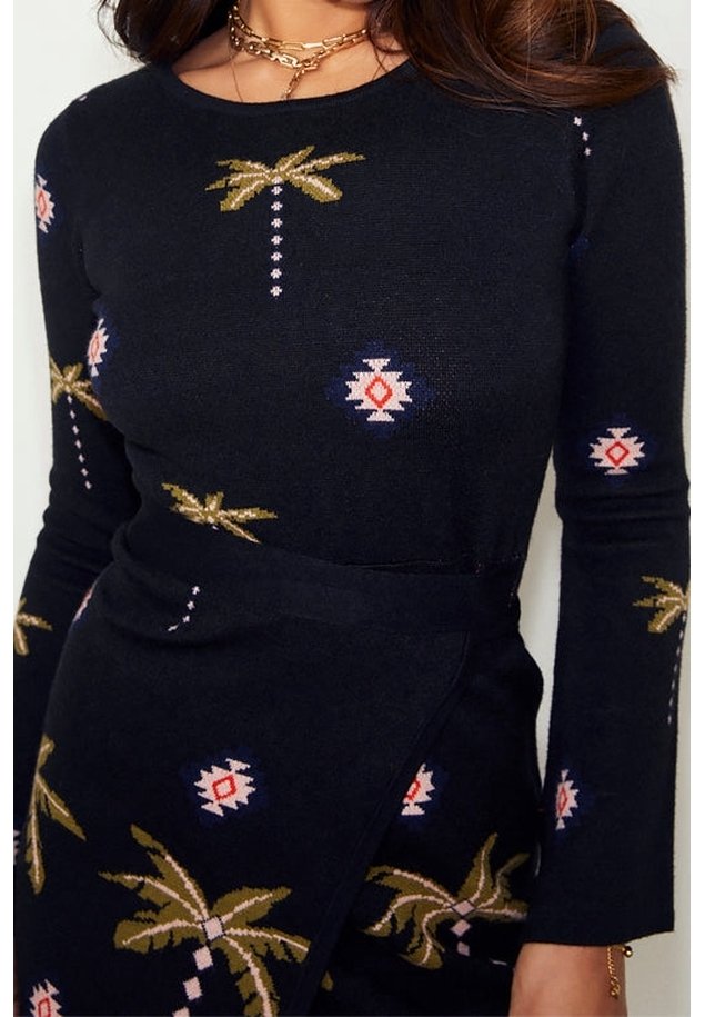 Femme portant une robe noire à manches longues avec un imprimé palmier et géométrique, accessoirisée avec des colliers en chaîne dorés superposés.