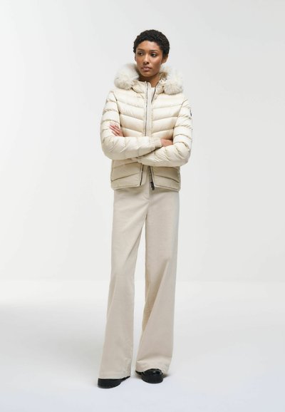 Femme portant une doudoune crème avec capuche en fourrure et pantalon beige à jambes larges, debout les bras croisés sur un fond blanc.