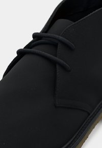 Μαύρο casual παπούτσι με στρογγυλεμένη μύτη, ραμμένες ραφές, δύο οπές για κορδόνια και μαύρα κορδόνια σε ανοιχτό φόντο.