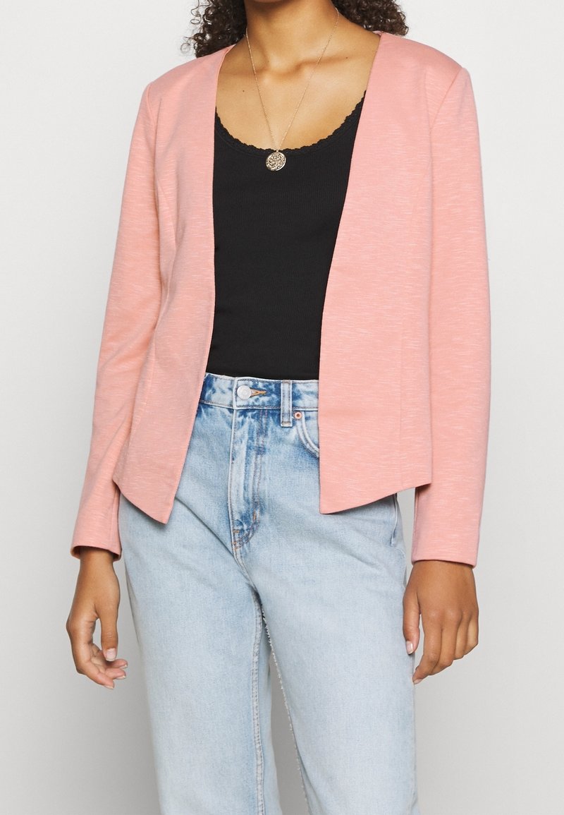 Femme portant un blazer rose clair ouvert sur un haut noir et un jean taille haute bleu clair, accessoirisée d'un petit collier pendentif rond.