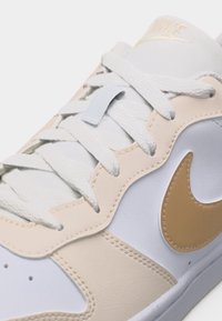 Κοντινή λήψη ενός μπεζ και λευκού αθλητικού παπουτσιού με λευκά κορδόνια και χρυσό σήμα Nike στο πλάι.