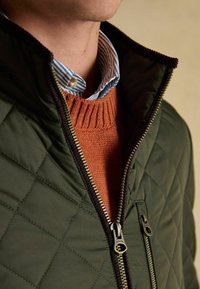 Mann trägt grüne gesteppte Jacke mit teilweise offenem Reißverschluss, orangefarbenen Strickpullover und blau-weiß gestreiftes Hemd mit Kragen.