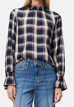 Kvinde iført en marineblå og beige ternet bluse med lange ærmer, med flæset høj hals og manchetter, stoppet ind i højtaljede blå jeans.