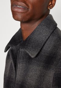 Närbild på en person som bär en mörkgrå ullkappa med en stor krage, silverfärgad hoop-örhänge och läppar delvis åtskilda.