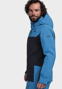 Blau- und schwarze Outdoor-Jacke mit Kapuze, die über ein schlankes Design, eine glatte Textur und ein Logo am Ärmel verfügt. Mehrere Taschen sind sichtbar.