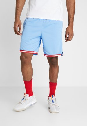 Homme portant un short de sport bleu clair avec des finitions rouges et blanches, des chaussettes rouges montantes et des baskets blanches avec des lacets bleus et orange, debout sur un sol blanc.