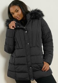 Svart pufferjacka med huva i pälsimitation, med kviltad design, knäppning och dragkedja, samt sidofickor. Slät textur.