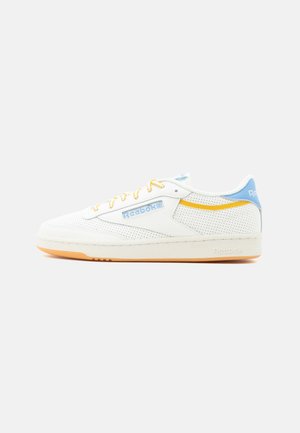 Witte lage Reebok-sneaker met geperforeerde zijkanten, gele en blauwe accenten, en een gumzool op een witte achtergrond.
