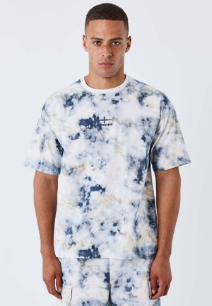 Man draagt bijpassend tie-dye blauw-wit T-shirt met korte mouwen en korte broek, staand tegen een effen witte achtergrond.