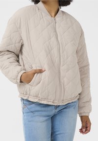 Person som bär en ljusbeige, vadderad bomberjacka med framdragkedja och sidofickor, i kombination med ljusblå jeans.