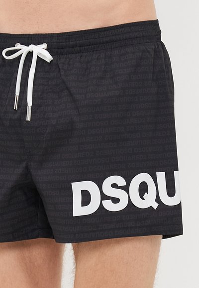 Schwarze Badehose mit weißem Kordelzug und großem weißen "DSQU"-Logo am linken Bein, getragen von einer Person mit heller Haut.