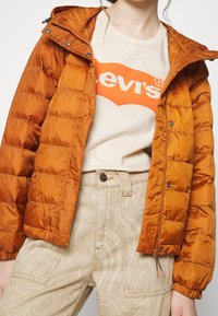 Veste matelassée orange avec capuche amovible et poignets élastiques, portée sur un t-shirt beige clair avec un logo rouge, associée à un pantalon beige à motifs.