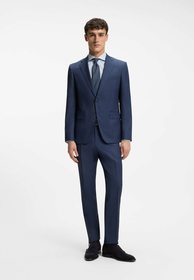 Homme debout portant un costume bleu marine ajusté avec une chemise bleu clair, une cravate bleu marine à pois, une ceinture noire et des chaussures noires sur fond blanc.