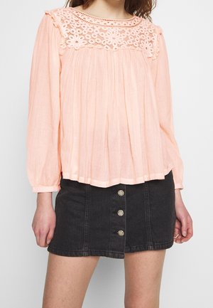 Vrouw die een perzikkleurige blouse met kanten inzetstuk en lange mouwen draagt, gecombineerd met een zwarte mini-rok van denim met knopen aan de voorkant.