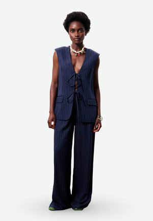 Vrouw draagt een marineblauw mouwloos vest met krijtstreep en wijde pijpen broek, gecombineerd met een schelpenketting en een zilveren armband, staand tegen een witte achtergrond.