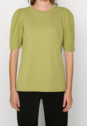 Femme portant un haut vert clair à manches courtes avec des épaules bouffantes et un pantalon noir, debout devant un fond uni.