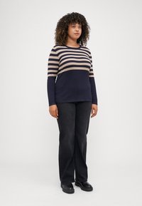 Haut à rayures bleu marine et beige à manches longues avec un col rond, associé à un pantalon large gris foncé et des chaussures à talons épais noires.