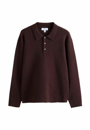 Mann lehnt drinnen an einer weißen Wand, trägt einen dunkelbraunen Pullover, helle Hosen und braune Stiefel, steht nahe einer Holztreppe.