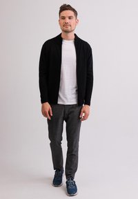 Musta vetoketjullinen takki valkoisen t-paidan päällä, yhdistettynä harmaisiin kuvioituihin housuihin ja sinisiin urheilulaihin. Malli seisoo yksinkertaista taustaa vasten.