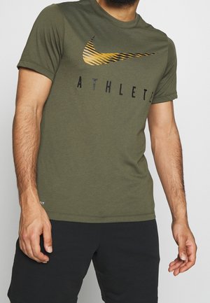 Mężczyzna w oliwkowej koszulce Nike z tygrysim paskiem na logo i napisem „ATHLETE”, w czarnych szortach, stojący na białym tle.