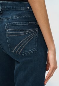 Κοντινό πλάνο πίσω τσέπης σκουρομπλε τζιν με καμπυλωτό ασημένιο ράψιμο και ένα χέρι που ακουμπάει δίπλα στον γοφό.