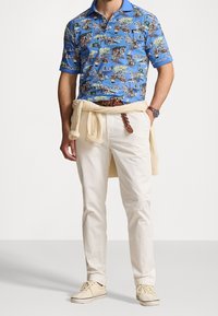 Lyhythihainen sininen polo, jossa on värikäs kuvio, valkoiset housut ja beige neule, joka on solmittu vyötärölle. Vaaleat tennarit täydentävät asua.