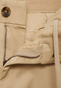 Bežna tkanina z gladko teksturo, z zadrgo in gumbom za zapiranje. Vključuje vrvico in šivane dodatke ob robovih.