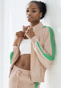 Light pink zipped jacket with green accents on sleeves. White sports bra and drawstring sweatpants visible. Smooth fabric texture.