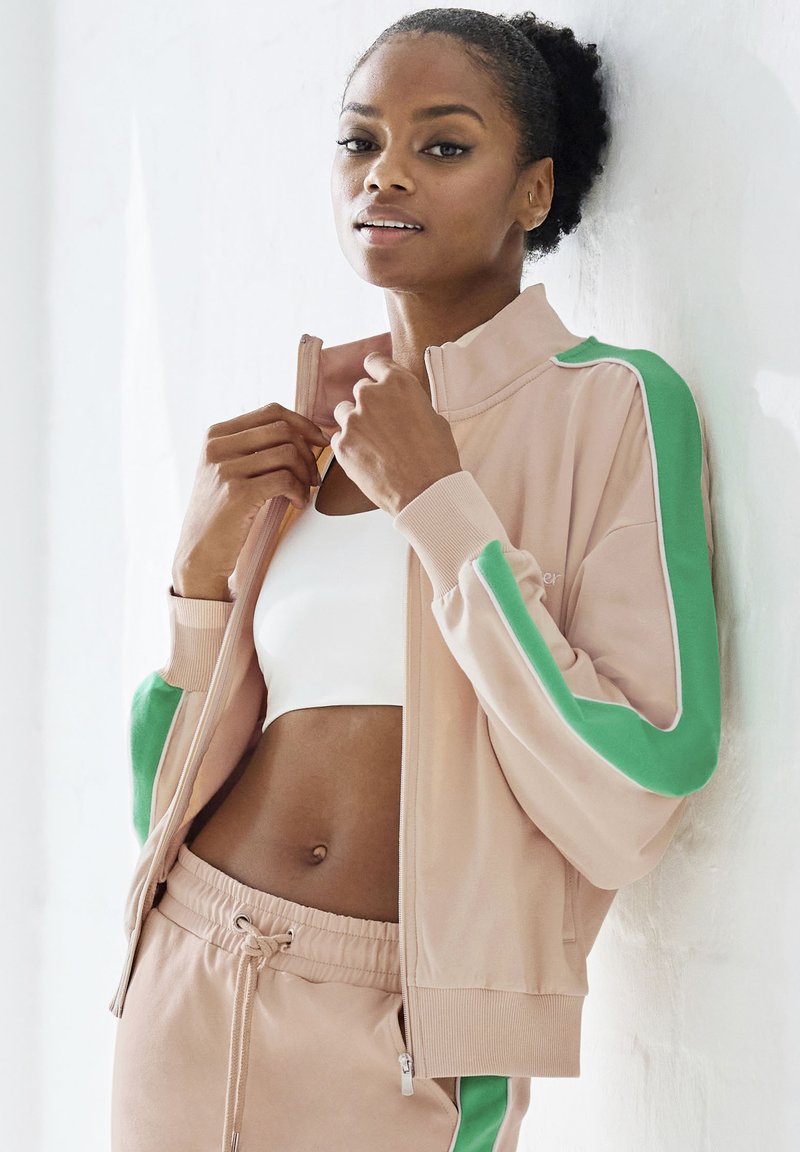 Light pink zipped jacket with green accents on sleeves. White sports bra and drawstring sweatpants visible. Smooth fabric texture.