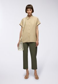 Camicia beige con bottoni e maniche corte, tessuto texturizzato, abbinata a pantaloni verdi oliva, borsa trasparente e sandali beige, su uno sfondo neutro.