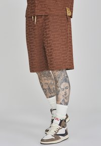 Braun gemusterte Shorts mit einem markanten Logo, kombiniert mit High-Top-Sneakers, weißen Crew-Socken und detaillierten Tattoos an den Beinen.