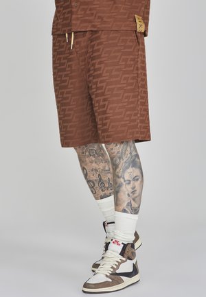 Pantaloni corti marroni a fantasia con un logo distintivo, abbinati a sneakers alte, calze bianche alla caviglia e tatuaggi dettagliati sulle gambe.
