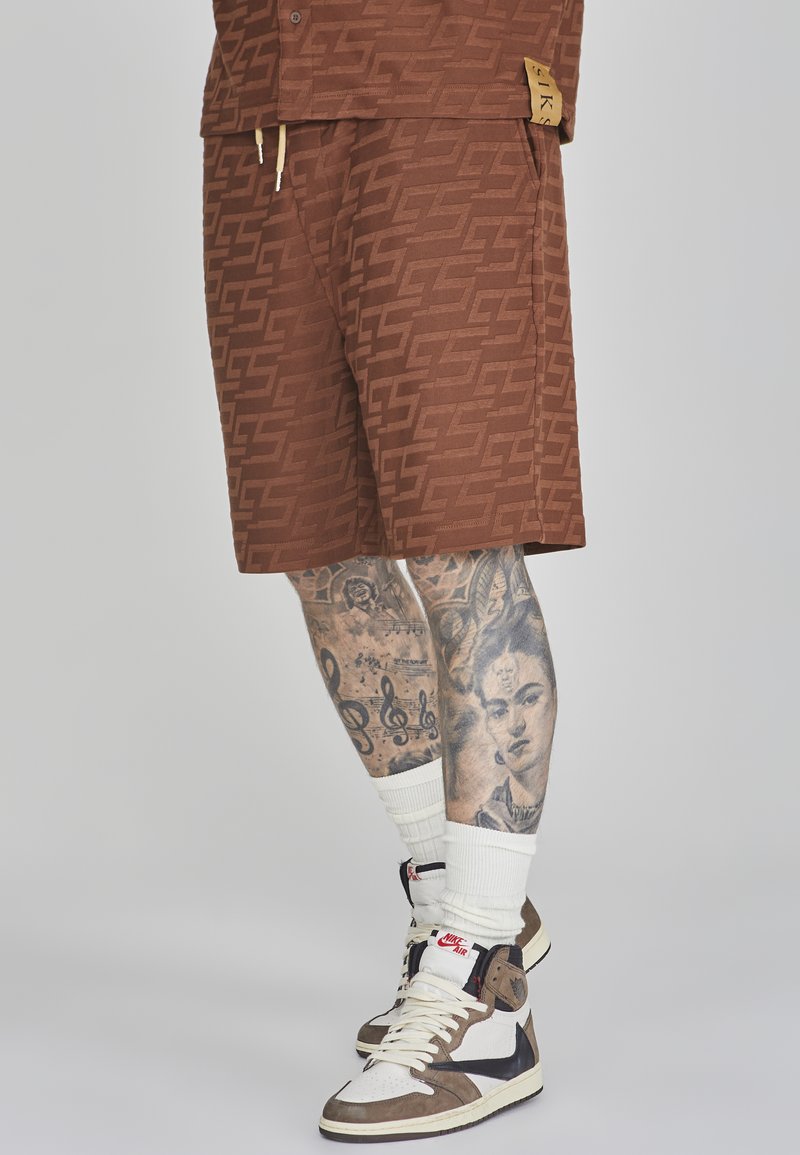 Braun gemusterte Shorts mit einem markanten Logo, kombiniert mit High-Top-Sneakers, weißen Crew-Socken und detaillierten Tattoos an den Beinen.