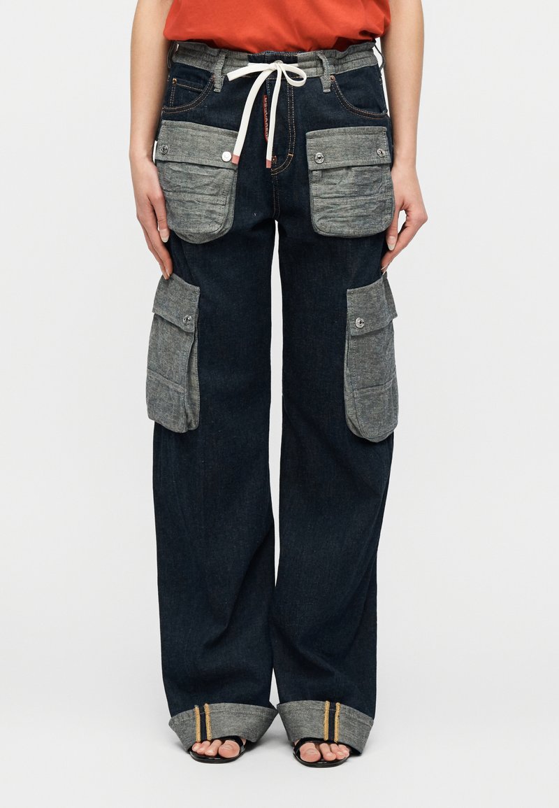 Γυναίκα που φορά σκούρο τζιν παντελόνι με φαρδιά μπατζάκια, μεγάλες γκρι τσέπες με κουμπιά και λευκό κορδόνι, σε συνδυασμό με μαύρα σανδάλια ανοικτά στα δάχτυλα.