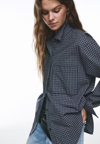 Košeľa s dlhým rukávom zapínaná na gombíky v námorníckej modrej kockovanej vzore, s náprsným vreckom a voľným strihom, skombinovaná so svetlomodrými džínsami.