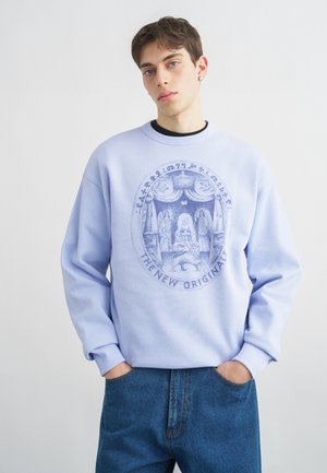 Noor mees lühikeste juustega, kandes helesinist dressipluusi, millel on detailne ovaalne embleem kujutiste, lõvi ja tekstiga "THE NEW ORIGINALS".