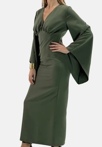Długa, oliwkowozielona sukienka z dekoltem w serek, podkreśloną talią i rozkloszowanymi rękawami, wykonana z gładkiej tkaniny. Na nadgarstku wyrazista bransoletka ze złotym wykończeniem.