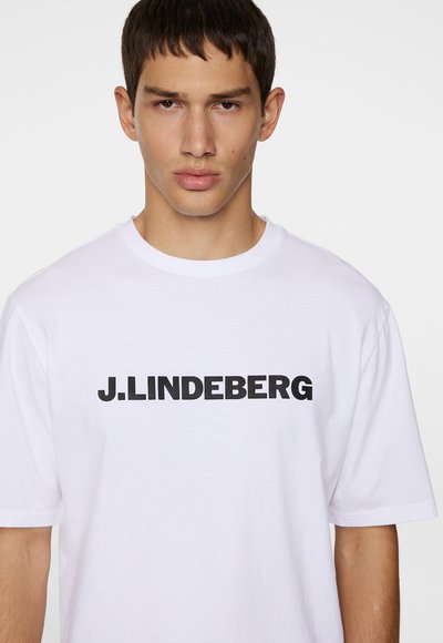 Valge puuvillane T-särk, millel rinnal on julge must logo "J.LINDEBERG". Klassikaline ümmargune kaelus ja lühikesed varrukad.