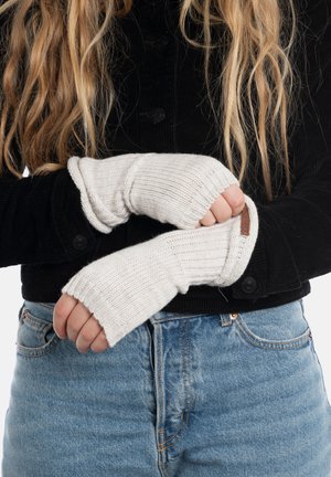 Gebreide vingerloze handschoenen in crèmekleur met ribbelstructuur en een leren label. Draagt het over een zwarte corduroy jas en blauwe spijkerbroek.