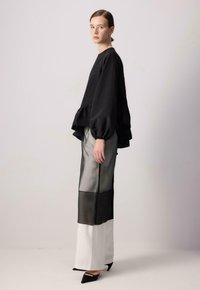 Melnas garās piedurknes blūze ar frillēm apakšmalā, savienota ar caurspīdīgām melnbaltām biksēm. Modelis valkā norādītās melnas kurpes ar augstiem papēžiem. Gluda tekstūra, brīvs piegriezums.