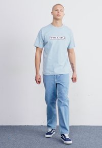 Camiseta de algodón azul claro con el logo negro de "VOLCOM", combinada con jeans azul claro y zapatillas marinas con acentos blancos.