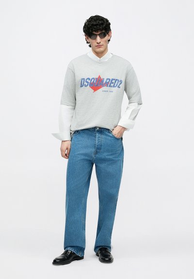 Graues Baumwoll-T-Shirt mit rotem und blauem "DSQUARED2"-Logo; geschichtete weiße Hemdärmel; blaue Jeans; schwarze Lederschuhe; stylische Sonnenbrille.