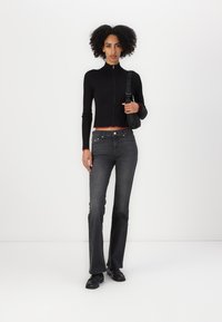 Black ribbed zip-up sweater with red accents, paired with flared dark grey jeans. Model holds a black shoulder bag. Smooth texture, fitted design.