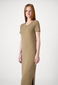 Robe longue de couleur kaki en tissu lisse. Elle a des manches courtes, un col rond et des fentes latérales pour faciliter les mouvements.