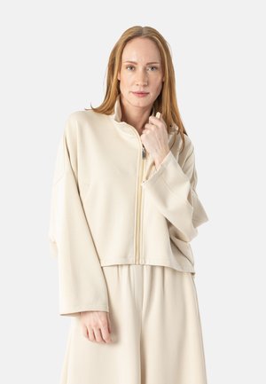 Kvinde med rødt hår iført løs beige lynlåsjakke og matchende vidde bukser, holder jakke kraven med den ene hånd.