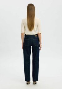 Mujer de pie de espaldas, vestida con una blusa corta de manga en color crema, jeans rectos de mezclilla azul oscuro, tacones beige, y con el cabello largo y lacio.