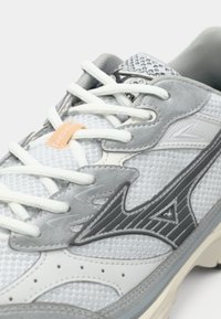 Gray and white athletic shoe featuring mesh texture, cushioned sole, and fabric laces. Notable gray accents and logo detail on the side.