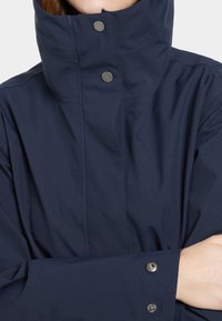 Marineblaue wasserfeste Jacke mit hohem Kragen, Druckknopfverschluss und glatter Textur mit sichtbaren Steppnähten.